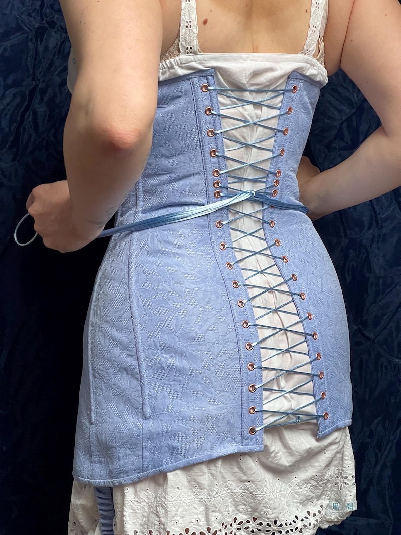 May include: A light blue corset with a floral pattern and copper-colored eyelets. The corset is laced up the back with blue ribbon. The garment is worn over a white top and skirt. The overall style is vintage.