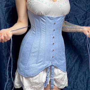 May include: A light blue corset with a floral pattern, featuring a row of small buttons down the front. The corset is paired with a white camisole and a white skirt with eyelet lace trim. The garment has suspender straps.