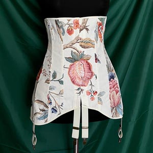 May include: A white corset with a floral pattern featuring pink, red, and blue flowers and green leaves. The corset has suspender straps and a structured design. The background is a dark green.