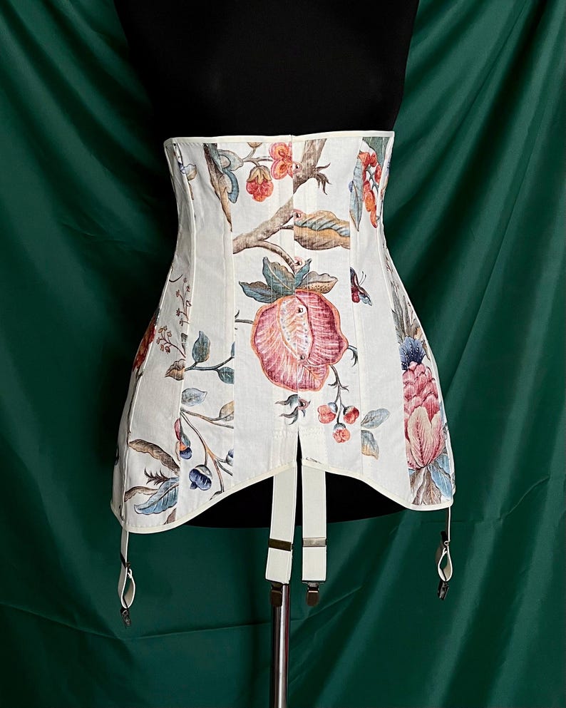 May include: A white corset with a floral pattern featuring pink, red, and blue flowers and green leaves. The corset has suspender straps and a structured design. The background is a dark green.