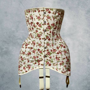 May include: A cream-colored corset with a floral pattern of red roses and green leaves. The corset has a button-up front, a structured silhouette, and suspender straps with metal clasps. The garment is designed to cinch the waist.