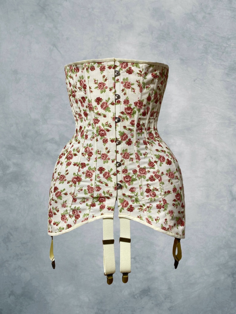 May include: A cream-colored corset with a floral pattern of red roses and green leaves. The corset has a button-up front, a structured silhouette, and suspender straps with metal clasps. The garment is designed to cinch the waist.
