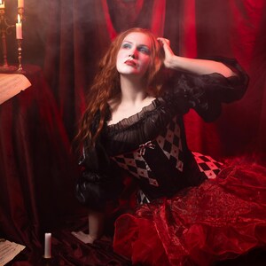 May include: A woman in a black and red costume with a diamond pattern bodice and a layered red skirt. She has red hair and is looking upward. The scene is set against a red backdrop with candles and sheet music.