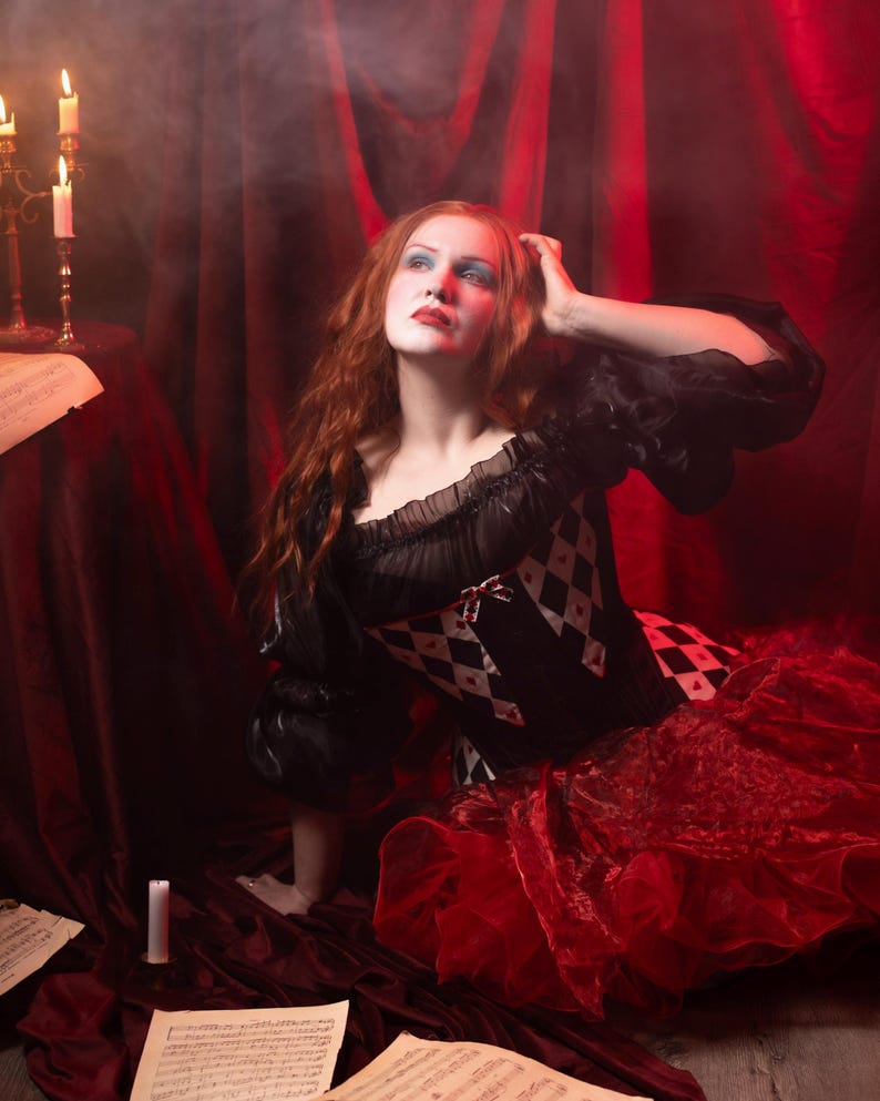 May include: A woman in a black and red costume with a diamond pattern bodice and a layered red skirt. She has red hair and is looking upward. The scene is set against a red backdrop with candles and sheet music.