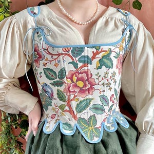Peut inclure: Un corset à motifs floraux sur fond crème, avec des fleurs et des feuilles rouges, roses et vertes. Le corset est bordé d'un liseré bleu clair et de liens, porté avec une blouse crème et une jupe verte. Le style général est vintage.