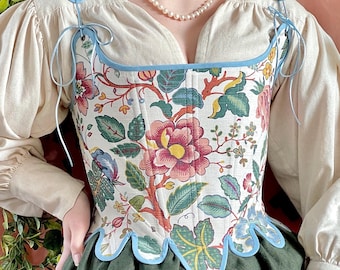 May include: A floral patterned corset with a cream background, featuring red, pink, and green flowers and leaves. The corset has a light blue trim and ties, and is paired with a cream-colored blouse and a green skirt. The overall style is vintage.