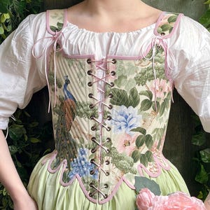 May include: A corset bodice featuring a cream-colored fabric with a print of peacocks and floral patterns in shades of green, blue, and pink. The garment has a front lace-up closure with metal grommets, pink trim, and shoulder straps with ties.