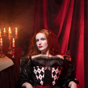 May include: A woman in a black and white corset with a red ruffled skirt sits in front of a red curtain. The corset features a diamond pattern with red hearts. Lit candles and sheet music are visible in the background.