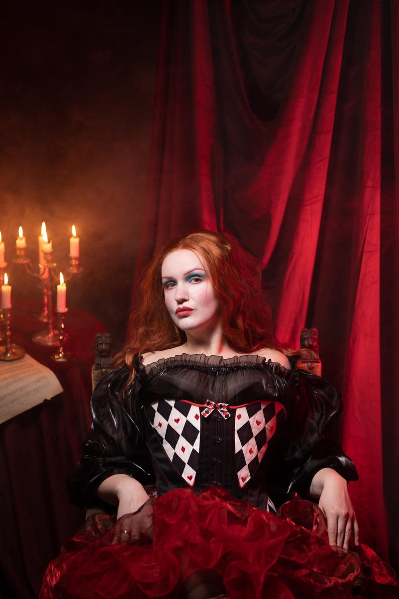 May include: A woman in a black and white corset with a red ruffled skirt sits in front of a red curtain. The corset features a diamond pattern with red hearts. Lit candles and sheet music are visible in the background.