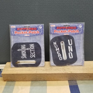 May include: Two zippered pocket patches, one with the text "Smoking Section" and the other with the text "Numero Uno". Both patches are black with white text and a silver zipper.