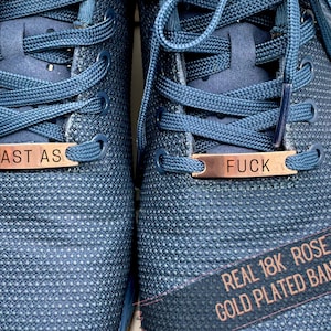 May include: Close-up of blue athletic shoes with matching laces. The shoes have rose gold-colored metal bands that read "FAST AS" and "FUCK". The text "REAL 18K ROSE GOLD PLATED BANDS" is also visible.