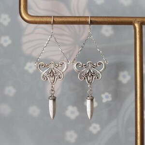 Peut inclure: Une paire de boucles d'oreilles argentées au design Art nouveau. Chaque boucle d'oreille présente un dessus décoratif avec une chaîne, un motif floral central et un pendentif pointu. Les boucles d'oreilles sont présentées sur un fond à motifs floraux.
