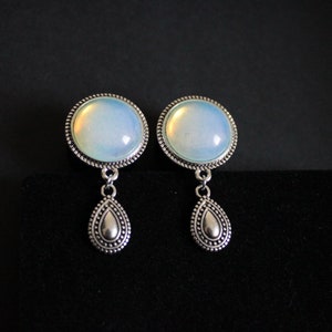 Può includere: Un paio di orecchini color argento con una pietra rotonda e opalescente incastonata in una cornice di perline. Ogni orecchino ha un pendente a forma di goccia. Sfondo nero.