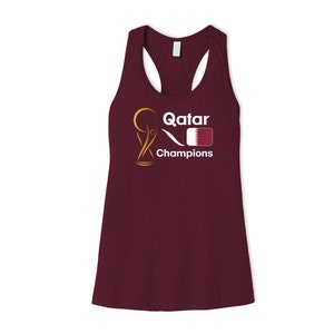 May include: A maroon tank top with a white graphic of a trophy, the word "Qatar", a stylized wave, and the flag of Qatar. The text "Champions" is below the graphic.