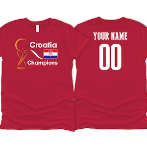 Puede incluir: Camiseta roja con un trofeo dorado, la palabra "Croacia", la bandera croata y la palabra "Campeones". La parte posterior de la camiseta tiene las palabras "Tu nombre" y el n&uacute;mero "00" en blanco.