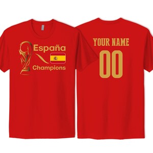 May include: A red short-sleeved t-shirt featuring a gold trophy graphic, the text Espa&ntilde;a Champions, and a Spanish flag on the front. The back displays the text YOUR NAME in gold above the gold numbers 00, centered on the red fabric.