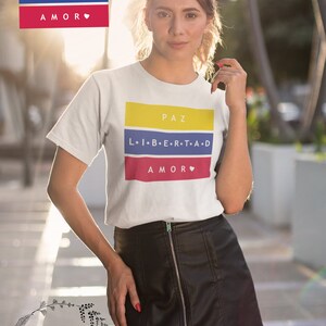 Pu&ograve; includere: Una maglietta bianca con un design grafico con i colori della bandiera colombiana. La grafica include le parole "PAZ", "LIBERTAD" e "AMOR" in spagnolo. La maglietta &egrave; indossata da una persona con una gonna di pelle nera.