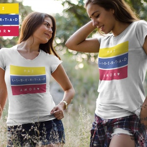 Pu&ograve; includere: Due donne indossano magliette bianche con un design grafico colorato. La grafica presenta le parole "PAZ", "LIBERTAD" e "AMOR" in giallo, blu e rosso, rispettivamente. Il design ricorda la bandiera colombiana.