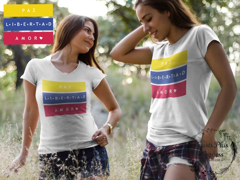 Pu&ograve; includere: Due donne indossano magliette bianche con un design grafico colorato. La grafica presenta le parole "PAZ", "LIBERTAD" e "AMOR" in giallo, blu e rosso, rispettivamente. Il design ricorda la bandiera colombiana.