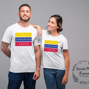 Pu&ograve; includere: Due magliette bianche con un design grafico con i colori della bandiera colombiana. La grafica include le parole "PAZ", "L-I-B-E-R-T-A-D" e "AMOR" in testo nero. Le magliette sono su uno sfondo grigio.