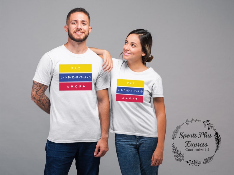 Pu&ograve; includere: Due magliette bianche con un design grafico con i colori della bandiera colombiana. La grafica include le parole "PAZ", "L-I-B-E-R-T-A-D" e "AMOR" in testo nero. Le magliette sono su uno sfondo grigio.