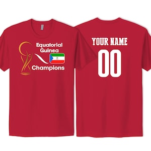 May include: Two red t-shirts, one with the text "Equatorial Guinea Champions" and a soccer trophy and flag, and the other with the text "YOUR NAME 00".