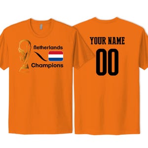 May include: An orange short-sleeved t-shirt featuring a gold trophy graphic, the text Netherlands Champions, and a Dutch flag on the front. The back displays the text YOUR NAME above the black numbers 00 in a bold, block-style font.