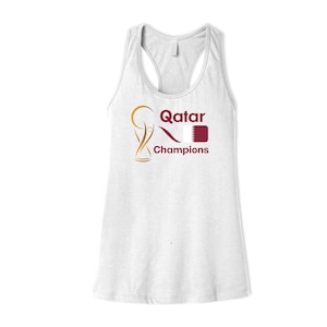 May include: White tank top with a gold and maroon graphic of a trophy, the word "Qatar", and a flag. The text "Champions" is below the graphic.
