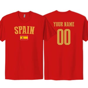May include: A red short-sleeved t-shirt showing the front and back views. The front features the word SPAIN in gold letters above the Spanish flag. The back displays the text YOUR NAME in gold above the gold numbers 00.