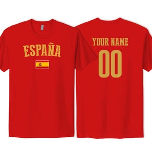 May include: A red short-sleeved t-shirt showing front and back views. The front features the text ESPA&Ntilde;A in gold above a small Spanish flag. The back displays the text YOUR NAME in gold above the gold numbers 00, centered on the red fabric.