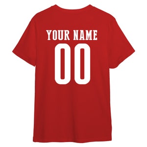 以下が含まれることがあります： 赤色のTシャツ。白で「YOUR NAME」の文字と数字の「00」がプリントされています。Tシャツは無地の赤色で、半袖です。