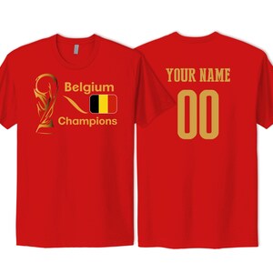 May include: A red short-sleeved t-shirt featuring a gold trophy graphic, the text Belgium Champions, and the Belgian flag on the front. The back displays the text YOUR NAME above the number 00 in a large gold font, centered on the fabric.
