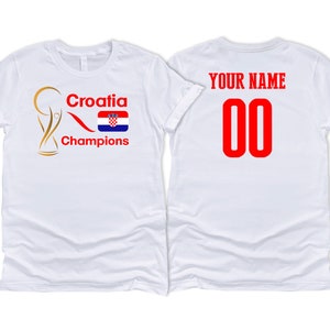 Puede incluir: Dos camisetas blancas, una con un trofeo dorado, el texto "Croatia Champions" y la bandera croata, y la otra con el texto "YOUR NAME" y el n&uacute;mero "00" en rojo.