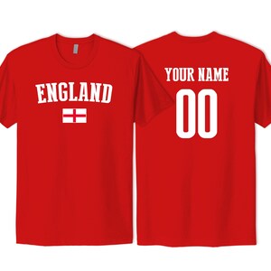 May include: A red short-sleeved t-shirt featuring the word ENGLAND in white capital letters above a white and red English flag on the front, and the text YOUR NAME above the number 00 in white on the back of the shirt.