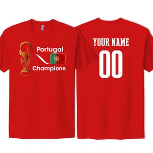 May include: A red short-sleeved t-shirt featuring a gold trophy graphic, the text Portugal Champions, and the Portugal flag on the front. The back displays the text YOUR NAME above the number 00 in large white font on the same red fabric.
