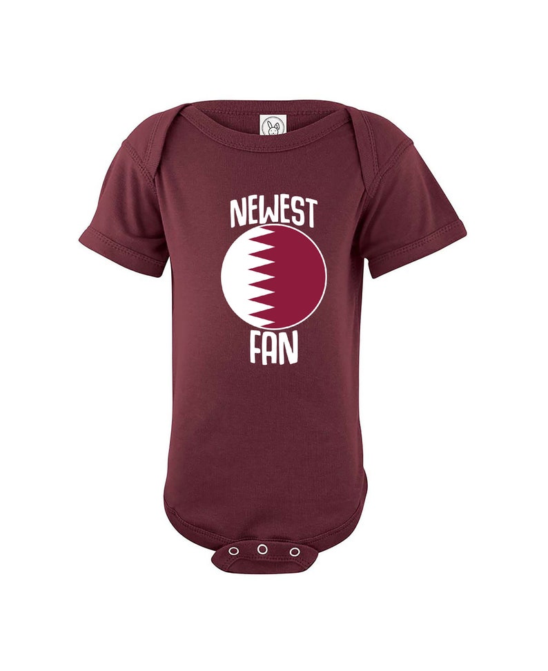 May include: A maroon baby onesie with a white circle featuring the flag of Qatar and the text "NEWEST FAN" in white.