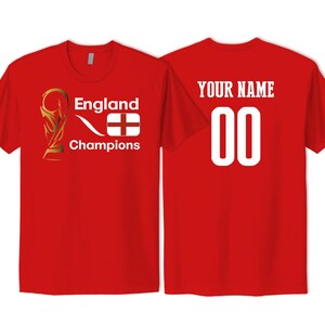 May include: A red short-sleeved t-shirt featuring a gold trophy graphic, the text England Champions, and an England flag on the front. The back of the shirt displays the text YOUR NAME above the number 00 in large white font.