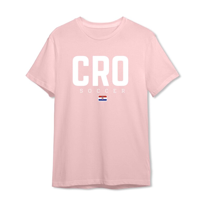 以下が含まれることがあります： 大きな白い文字で「CRO」と書かれた、明るいピンク色のTシャツ。「SOCCER」の文字が下にあり、その下にクロアチアの国旗が描かれています。クルーネックのTシャツです。