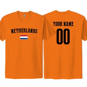 May include: An orange short-sleeved t-shirt featuring the text NETHERLANDS and a small Dutch flag on the front. The back of the shirt displays the text YOUR NAME above the large black numbers 00 in a bold, athletic-style font.