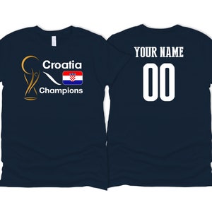 Puede incluir: Camiseta azul marino con un trofeo dorado, el texto "Croatia Champions" y la bandera croata. La parte posterior de la camiseta tiene el texto "YOUR NAME" y el n&uacute;mero "00".