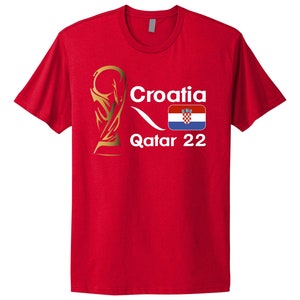 May include: Red t-shirt with a white graphic of a gold trophy, the word "Croatia", the Croatian flag, and the text "Qatar 22".