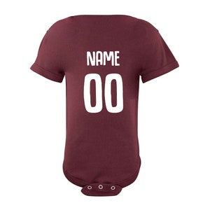 May include: Burgundy baby bodysuit with white text that says "NAME 00" on the back.