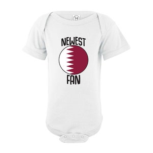 May include: White baby onesie with a red and white circle featuring the flag of Qatar. The text "NEWEST FAN" is printed above and below the circle.