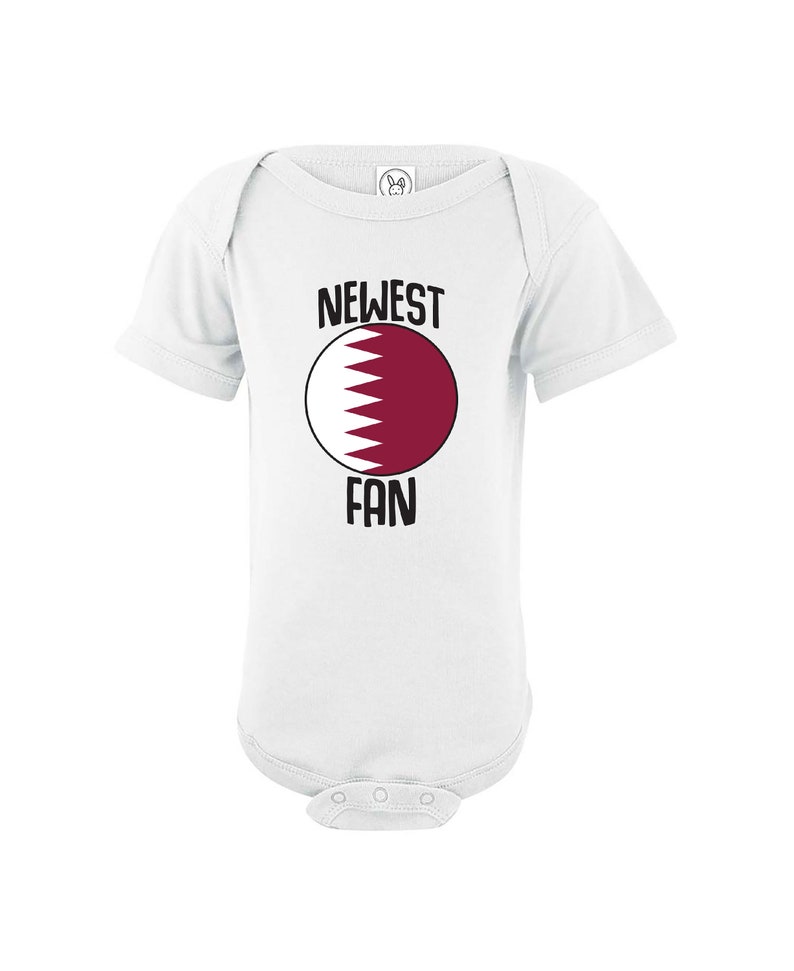 May include: White baby onesie with a red and white circle featuring the flag of Qatar. The text "NEWEST FAN" is printed above and below the circle.
