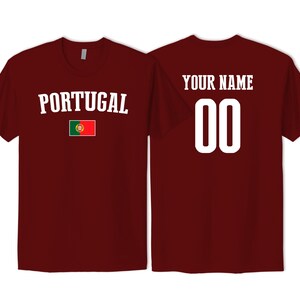 May include: A maroon short-sleeved t-shirt featuring the word PORTUGAL in white text above the Portugal national flag on the front, and the text YOUR NAME above the number 00 in white on the back of the garment.
