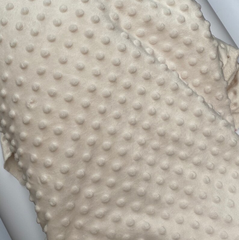 May include: Close-up of a cream-coloured fabric with a textured, raised dot pattern. The fabric is soft and plush.