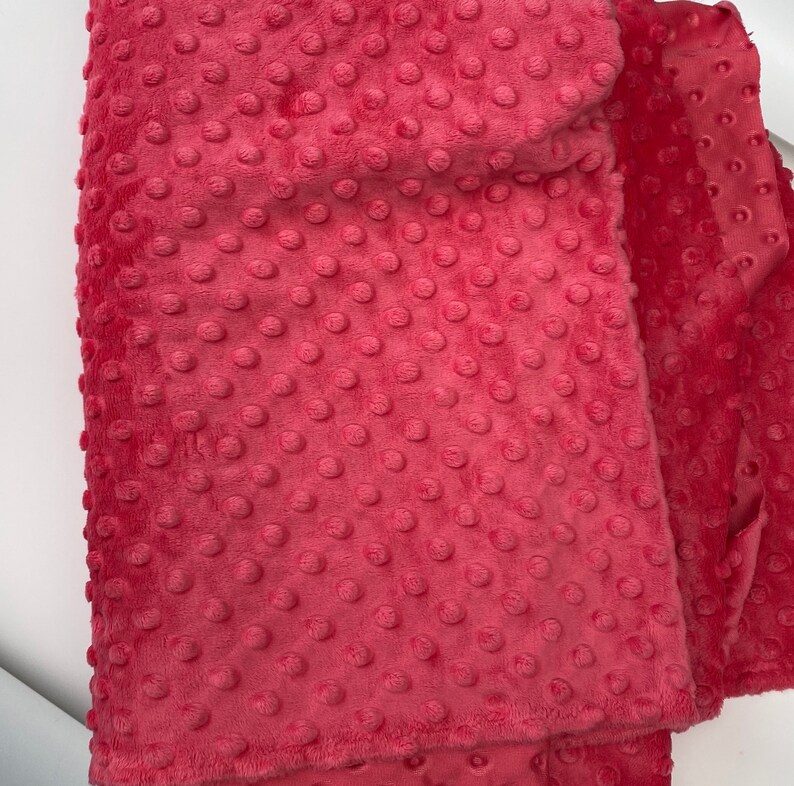 May include: A close-up of a pink minky fabric with a textured dot pattern. The fabric is soft and plush, ideal for blankets, baby clothes, and other crafts.