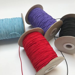 May include: Four spools of elastic cord in different colors: light blue, purple, black, and red. The spools are made of cardboard and have a hole in the center for dispensing the cord.