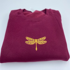 Può includere: Una felpa bordeaux piegata con un ricamo dorato di una libellula sul davanti. La libellula è dettagliata con motivi intricati. La felpa è realizzata in un materiale morbido.