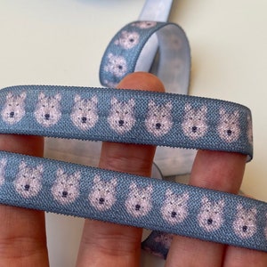 Puede incluir: Primer plano de una cinta elástica gris con un patrón repetido de caras de perros rosas. La cinta está doblada por la mitad y sostenida por un dedo.